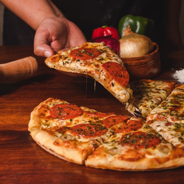 A hand lifts a slice of Angelo's pepperoni pizza with melted cheese, on a wooden table.